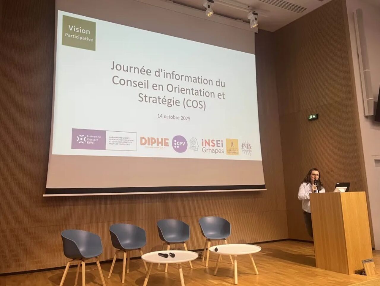 Caroline Pigeon ouvre la journée d'information du Conseil en Orientation et Stratégie, debout devant un pupitre, elle a dans son ods un vidéoprojecteur qui contient le nom de la journée et les logos des partenaires.